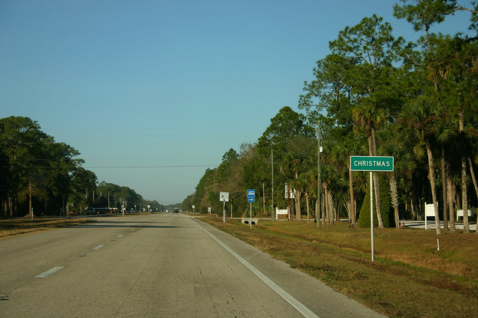 A green highway sign reading CHRISTMAS beside a wide, almost empty two-lane highway lined with tall pines and cabbage palms under a big blue sky.