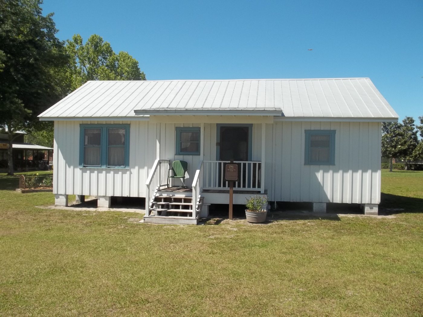 A small white board-and-batten house with a metal roof and a short covered porch, raised on blocks, standing on a wide mowed lawn with other historic buildings behind it.