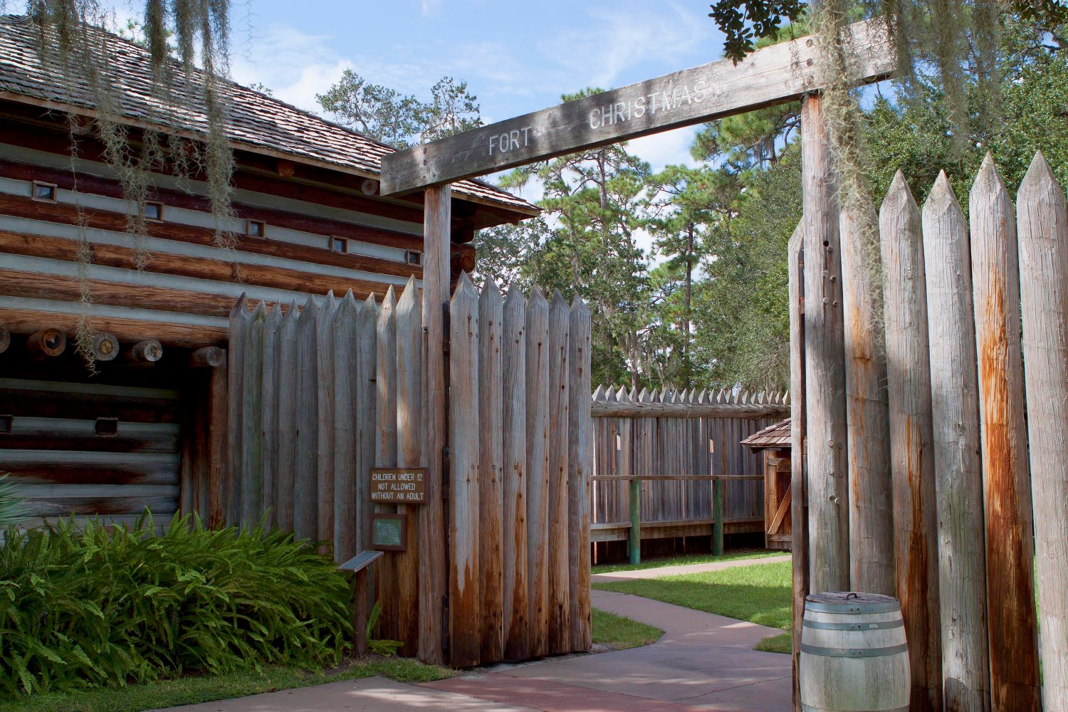 A log palisade fort gate with pointed upright timbers and a crossbeam carved with the words FORT CHRISTMAS, a shingled blockhouse to the left and Spanish moss hanging overhead.
