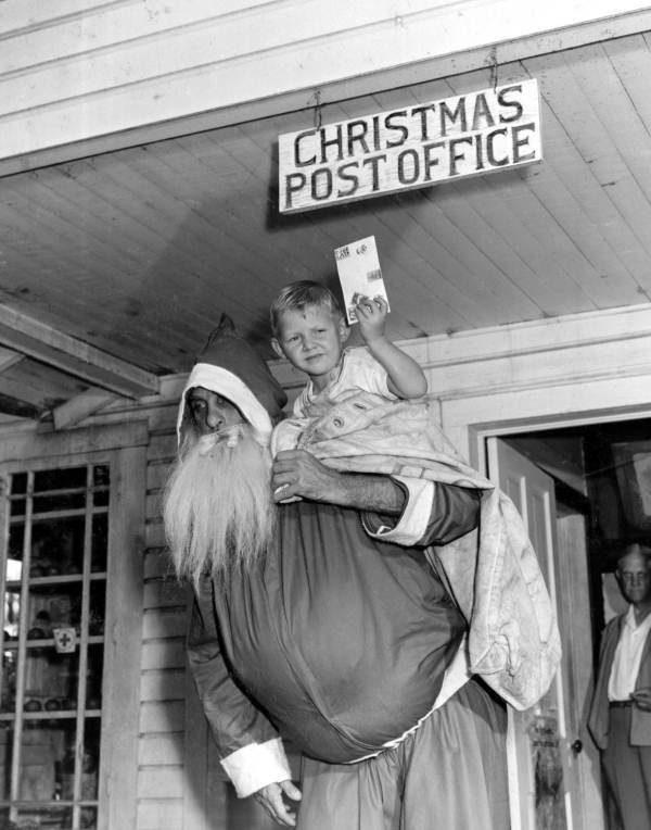 A 1947 black-and-white photo of a bearded Santa Claus carrying a small boy on his shoulder under a hand-painted sign reading CHRISTMAS POST OFFICE, the boy holding up a greeting card.