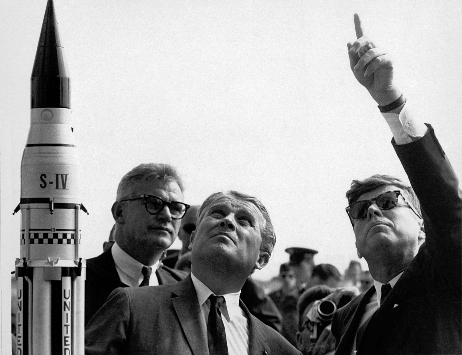 President John F. Kennedy and a group of men in suits standing outdoors at Cape Canaveral while Wernher von Braun gestures toward a rocket, pointing something out.