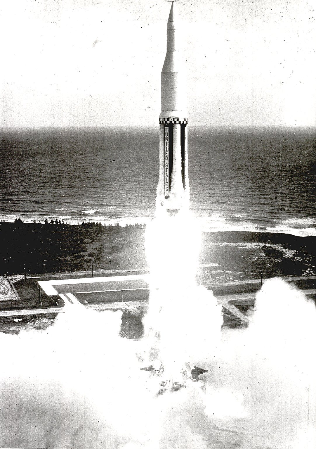 A tall Saturn rocket lifting off a coastal launch pad on a thick column of smoke and flame, gantry towers beside it.