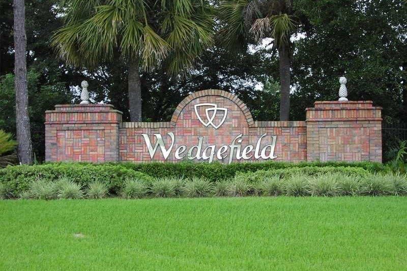 A low brick-and-stucco monument sign reading WEDGEFIELD at a neighborhood entrance, with manicured plantings and tall pines behind it under a blue sky.